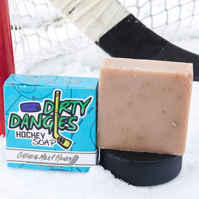 A brown bar of dirty dangles hockey soap sits on a hockey puck in the snow with a hockey stick and a hockey goal. Oatmeal Milk and Honey Scent