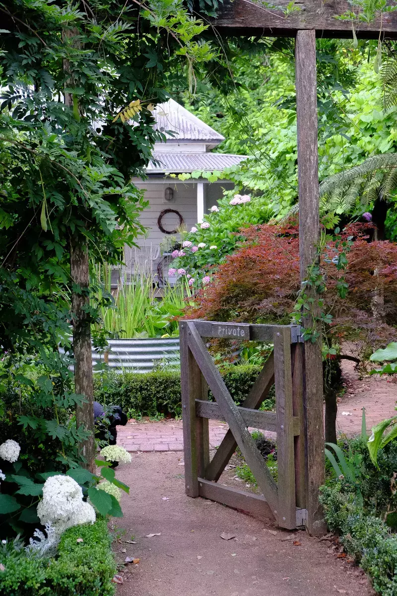 Rustic timber garden gate marked “Private,” set beneath a vine-covered pergola along a winding gravel path, surrounded by lush cottage garden plants and greenery with a country homestead in the background.