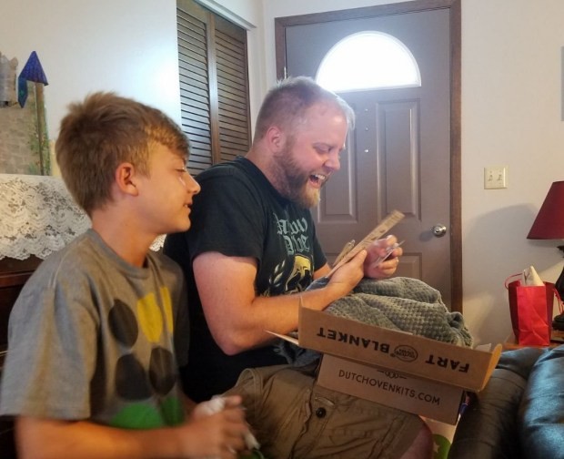 A man opening up a green dutch oven kits fart blanket gift box with his son and laughing