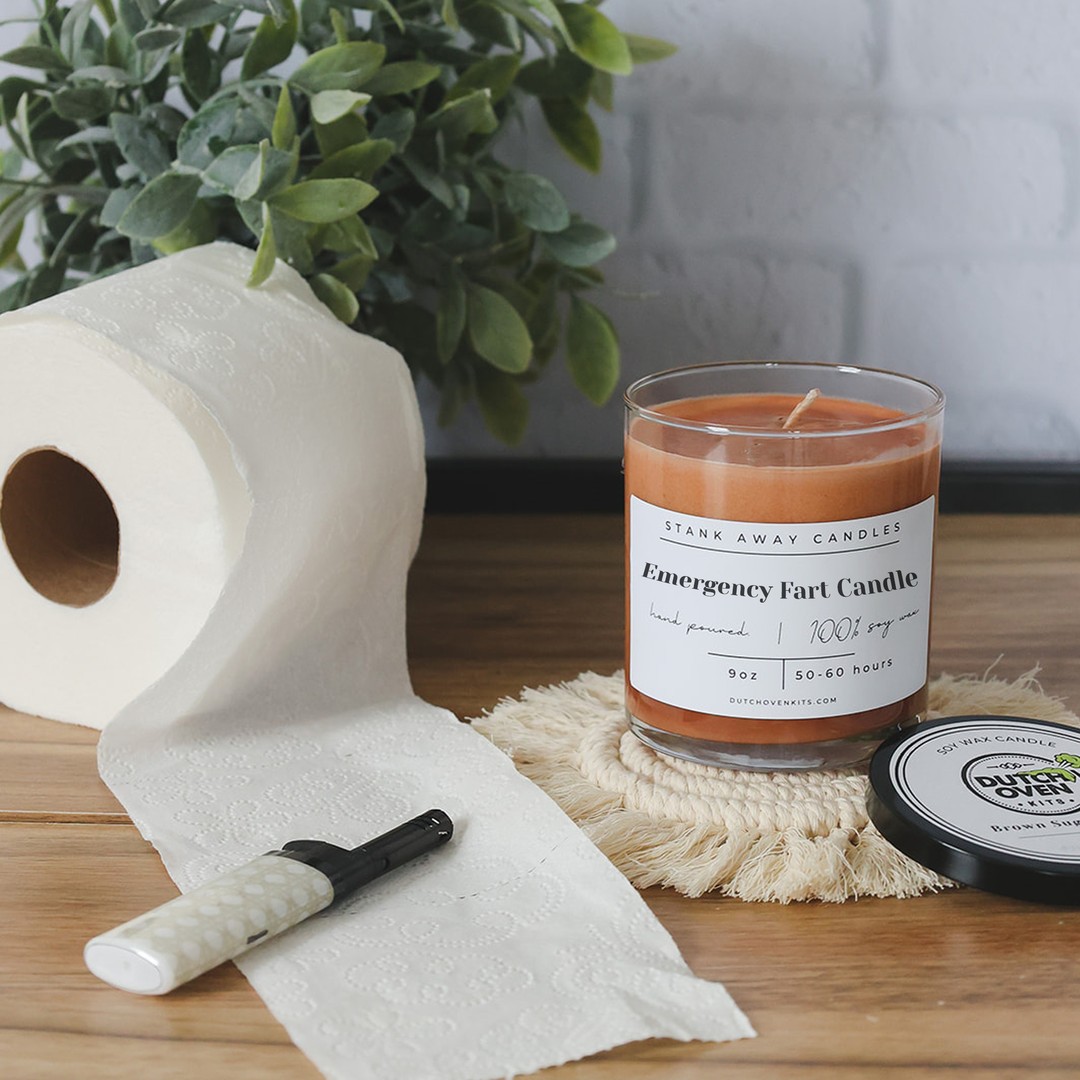 A brown candle sitting on a wood table with a roll of toilet paper and a lighter. Emergency fart candle