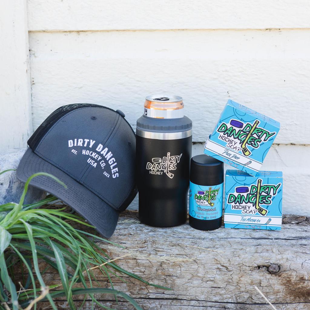 2 bars of hockey soap, a black 2 in 1 drink tumbler can holder, a stick of dirty dangles natural deodorant and a dirty dangles hockey co. hat outside on a piece of wood.