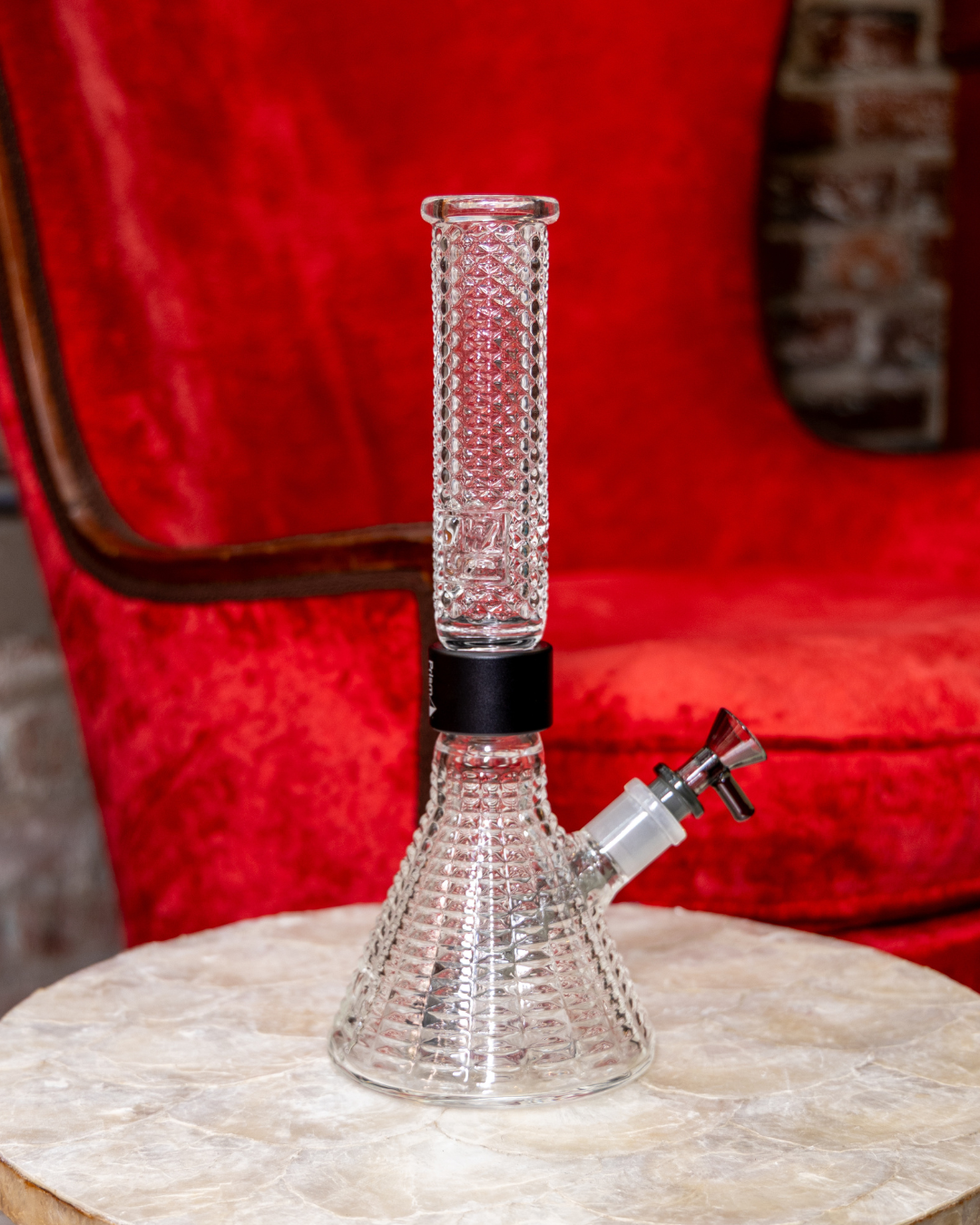 The clear Prism x Jane West Twenties MINI Beaker Bong displayed on a luxurious white marble table. The background features a striking red velvet chair and exposed red brick, highlighting the bong's role as an elegant centerpiece and accentuating the sophisticated, textural gridded diamond design.