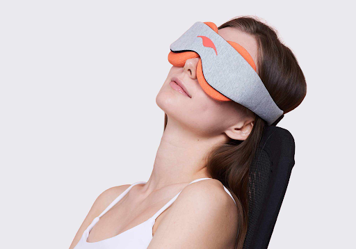 A girl reclining on her chair wearing a sleep mask with orange warming eye cups.