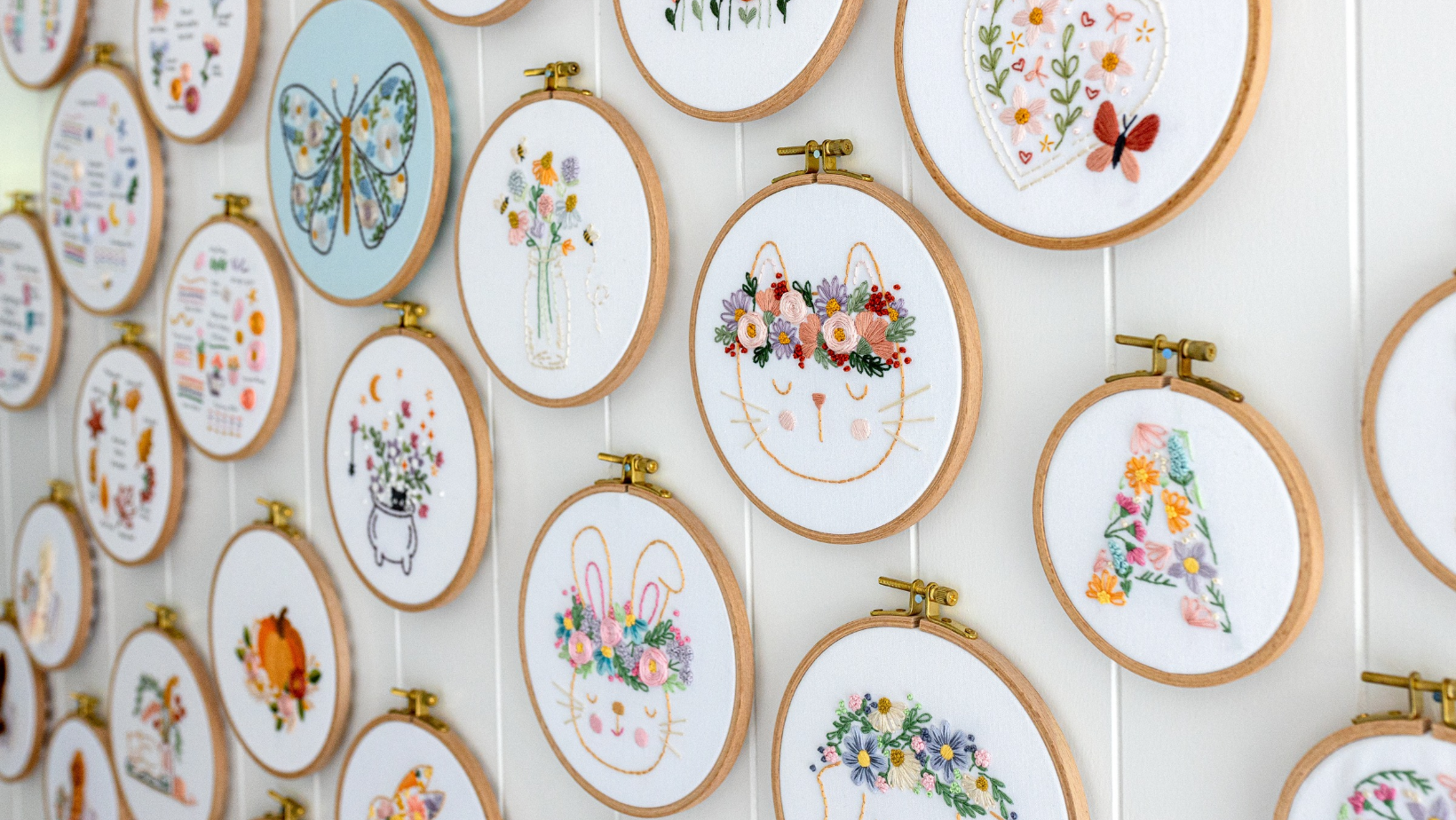 A wall display of dozens of finished embroidery hoops, including florals, a butterfly, a bunny and a cat with a flower crown.