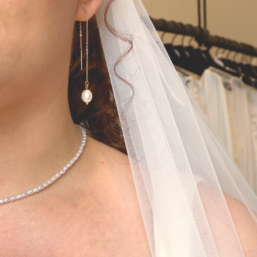 A woman wearing a white veil, a gold threader earring with a pearl drop and a pearl necklace.