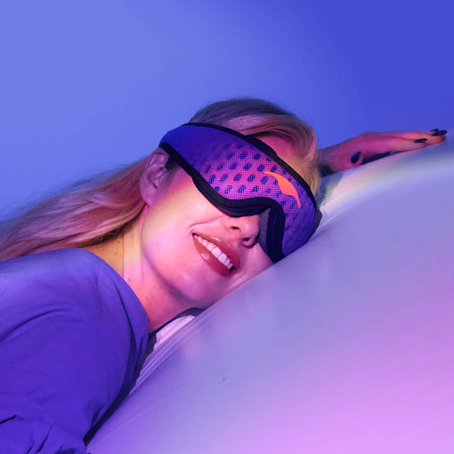 A smiling blonde girl sleeping on her stomach wearing a blue mesh. sleep mask