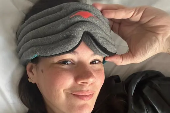 A smiling woman wearing a gray weighted eye mask that provides gentle pressure.