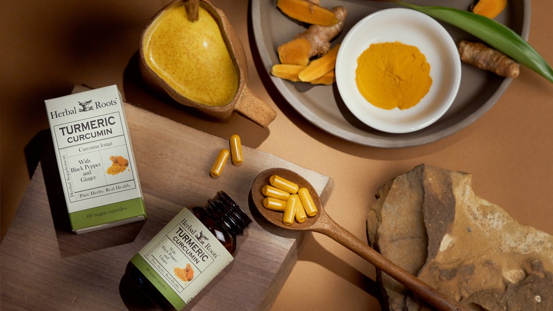 aerial view of a bottle of herbal roots turmeric supplement on its side with a cutting board next to it that has a wooden spoon with turmeric capsules in it and a mug of orange drink. To the left of the bottle is a ceramic plate with a small bowl of turmeric powder in it and a small pile of chopped turmeric root in front of it.