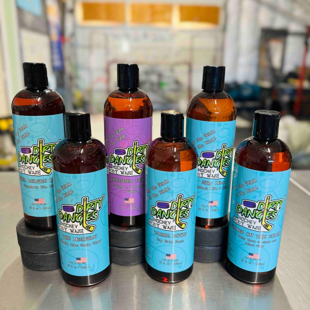 6 bottles of dirty dangles hockey body wash on a table with hockey nets in the background