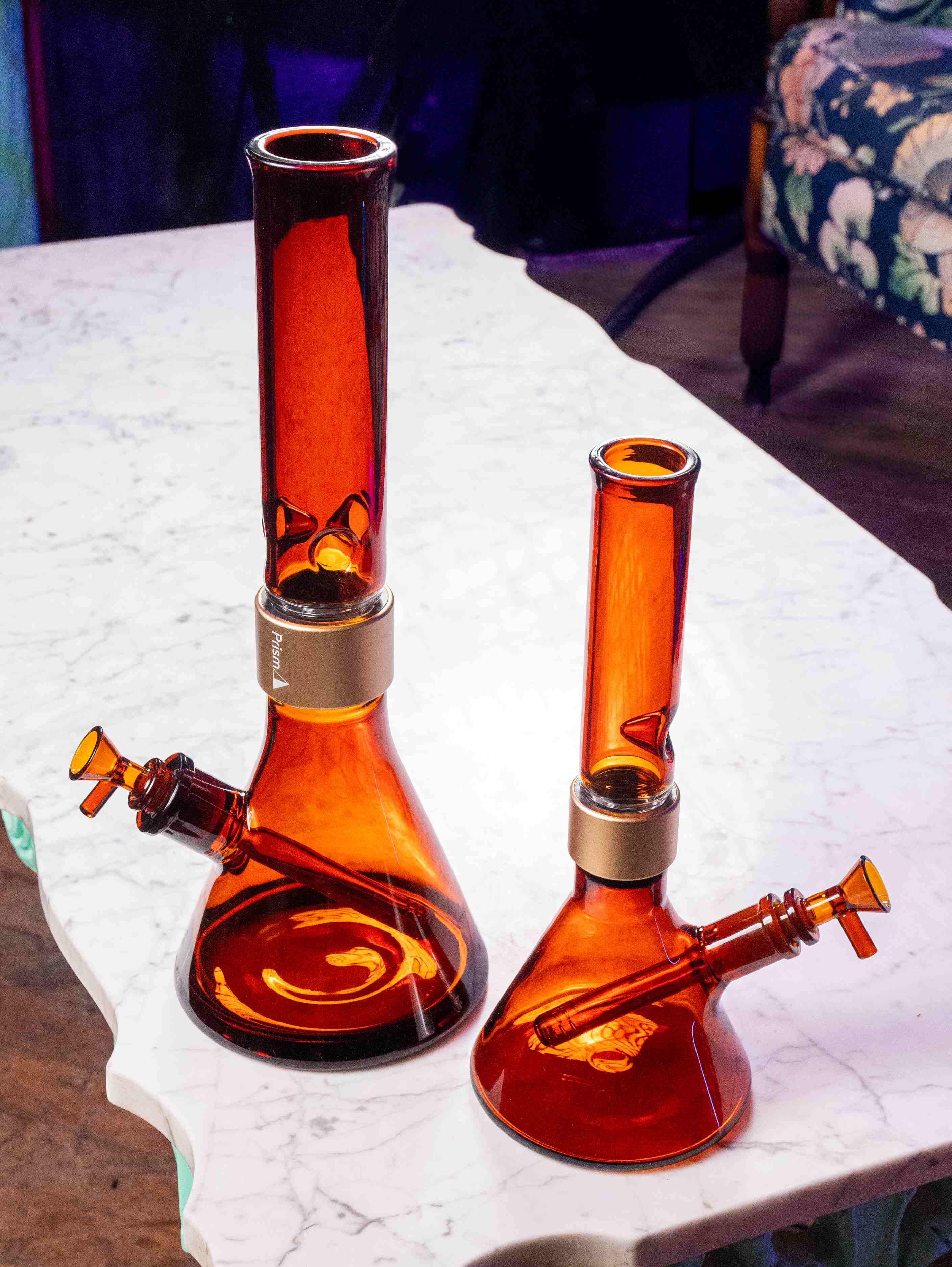 A side-by-side comparison of the Prism Amber Monochrome setups in Classic (left) and MINI (right) sizes. Both modular bongs feature amber beaker bases, matching amber bowls, downstems, and mouthpieces, all secured with signature gold halo connectors.