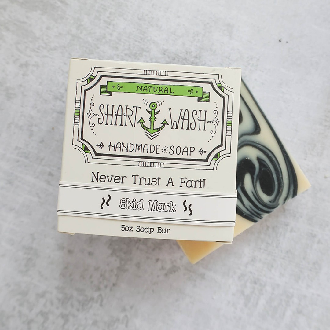 Picture of a box of Shart Wash Natural Handmade Bar Soap Skid Mark scent sitting on a black and white bar of soap with a wood background