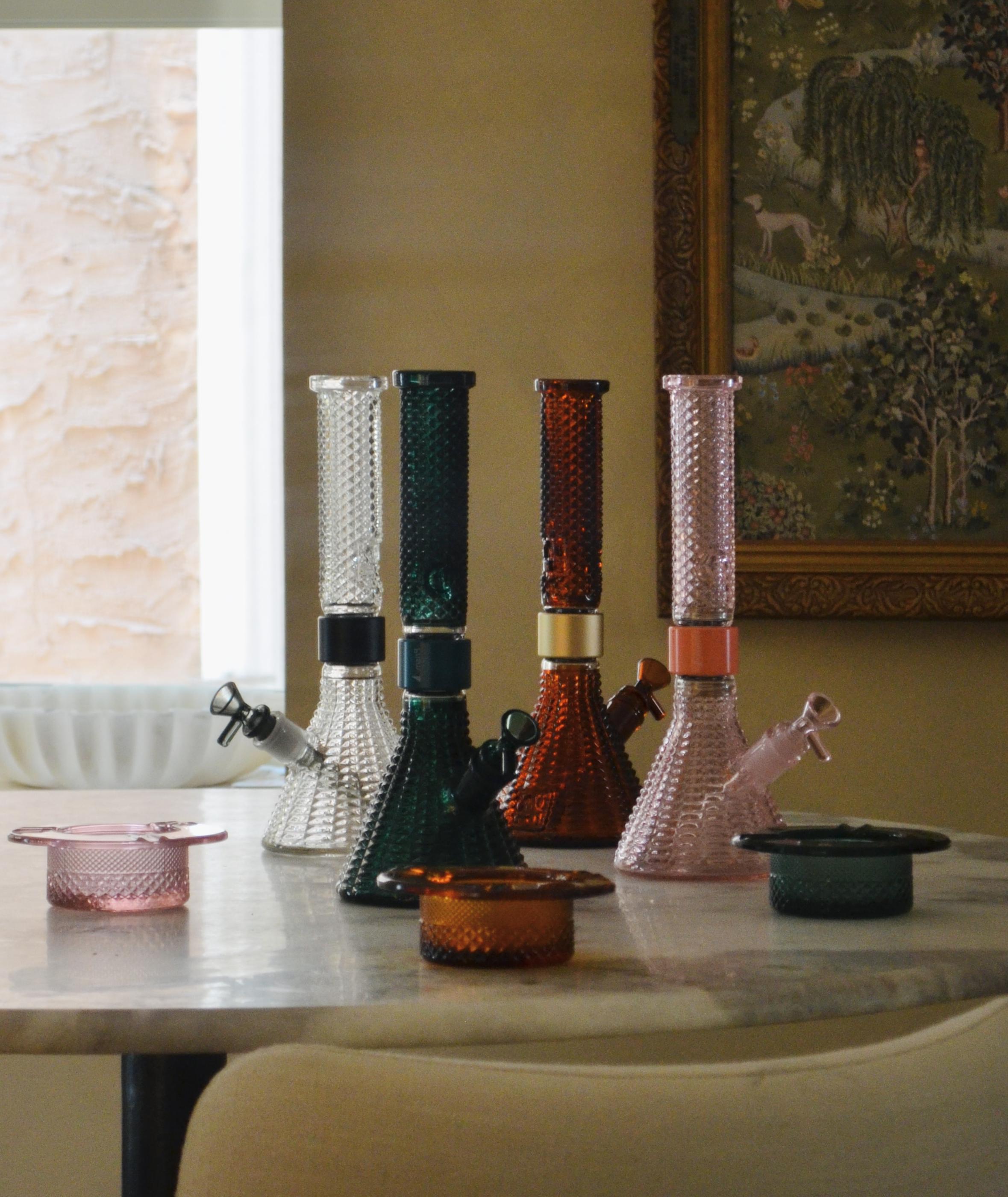 The Prism x Jane West Twenties MINI Beaker Bongs, in clear, teal, vintage amber, and pink lemonade colors in art Deco-inspired textured glass style on table lifestyle shot with matching ashtrays in pink, amber, and teal.