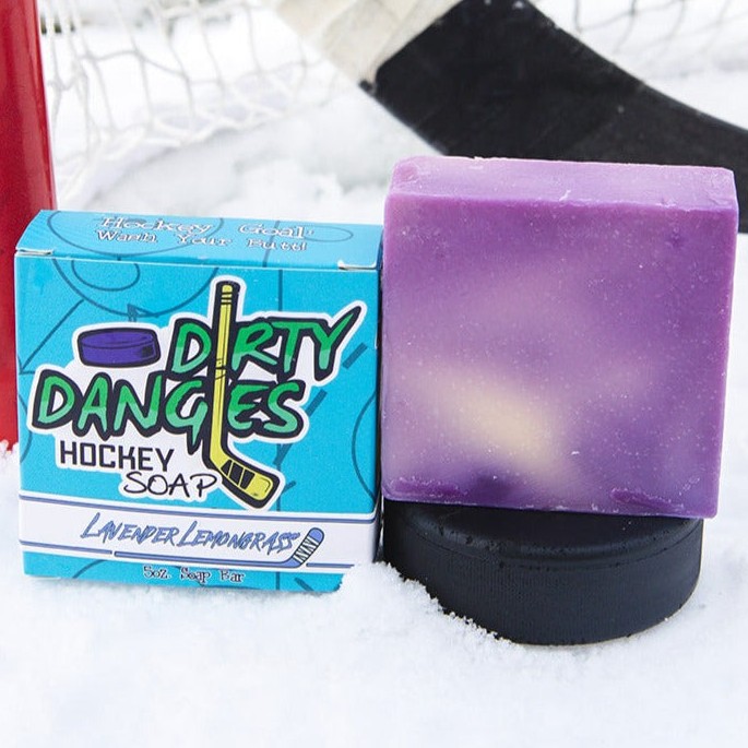 A purple and yellow bar of dirty dangles hockey soap sits on a hockey puck in the snow with a hockey stick and a hockey goal. Lavender Lemongrass Scent