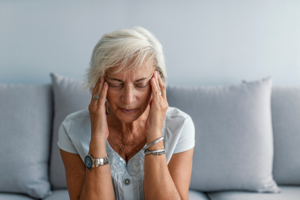 Senior woman sitting on couch, having a migraine. Gray background.