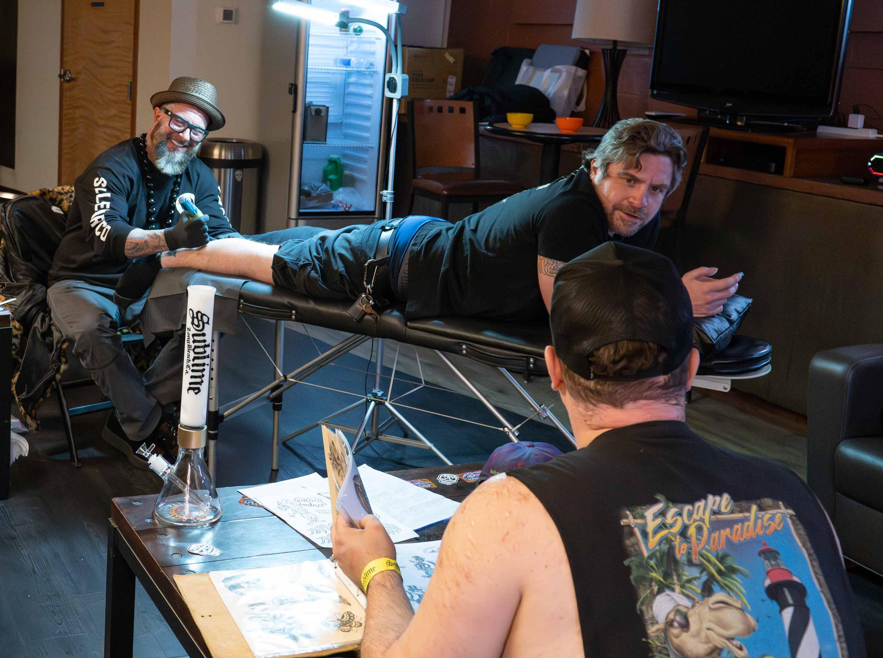 A Sublime x Prism official modular bong sits in the Red Rocks Amphitheatre greenroom while a fan gets the iconic Opie Ortiz Sublime Sun logo tattooed on his calf. Sublime band members look on, capturing the intersection of tattoo culture and the official glass collection backstage.