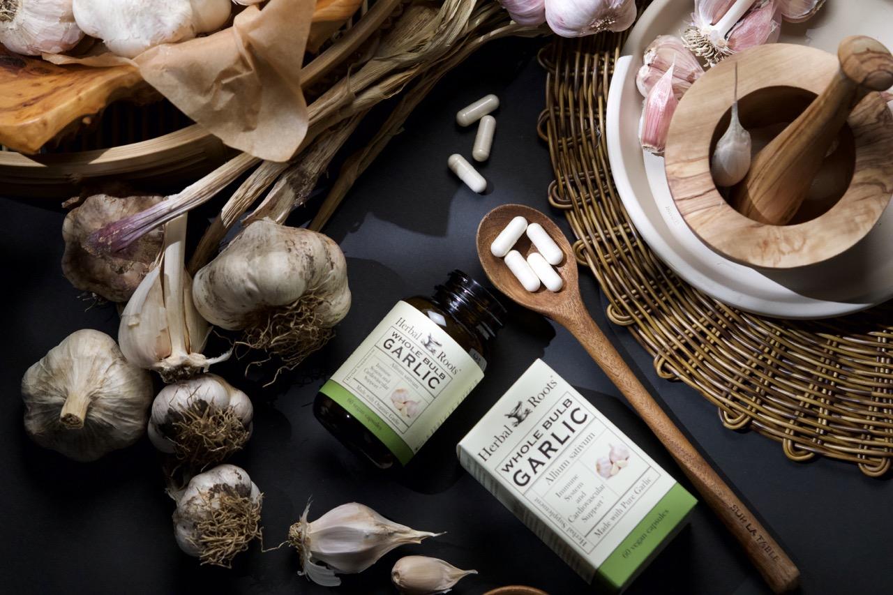 Ariel view of a bottle and box of Herbal Roots garlic supplement laying on their sides. There are whole bulbs of garlic, a wooden spoon with garlic capsules in it , a wood mortar and pestle on a white plate on a wicker placemat.