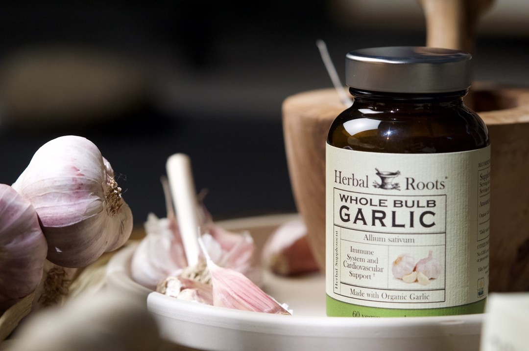 Bottle of Herbal Roots garlic with a wood mortar and pestle behind it. The bottle is sitting on a white plate with raw garlic next to it.