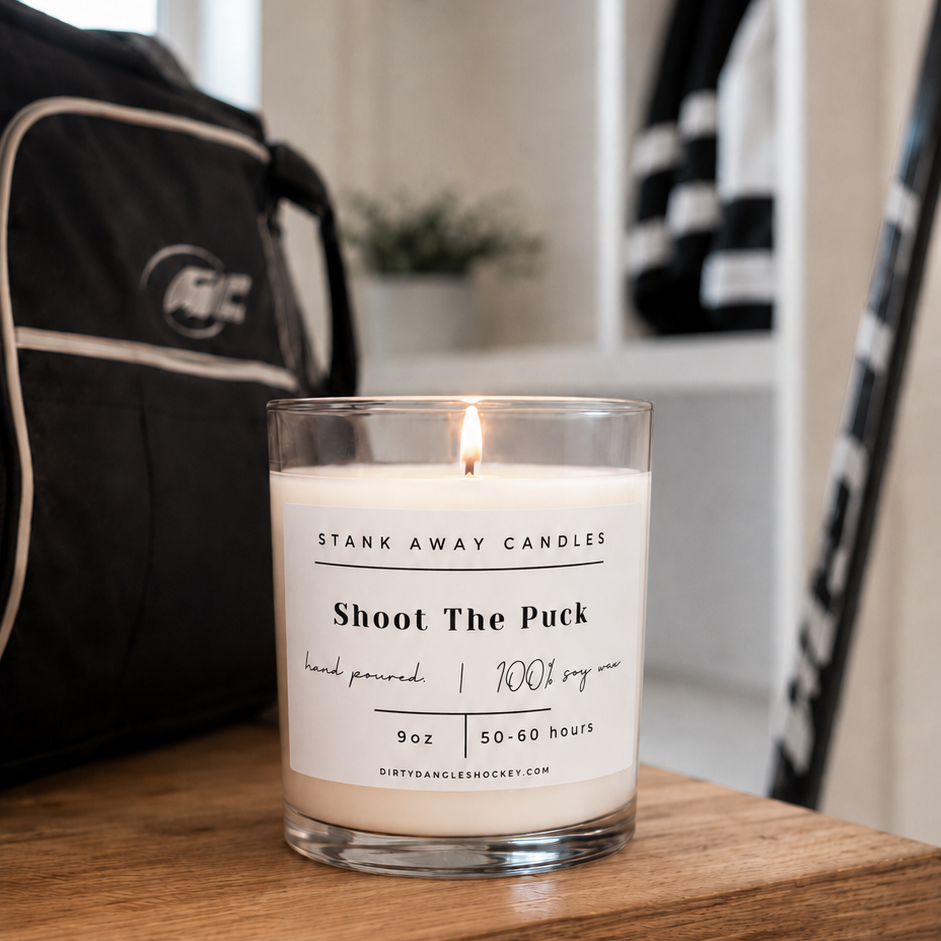 A white scented hockey candle on a wooden bench with hockey gear. Shoot the puck