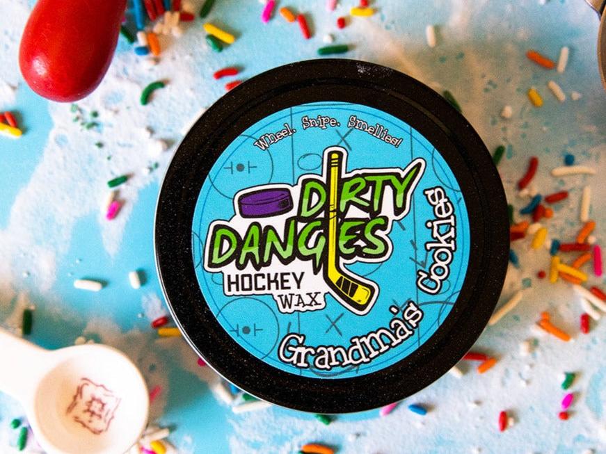 A tin of Dirty Dangles Hockey Stick Wax Grandma's Cookies Scent sits on a blue background surrounded by sprinkles and baking supplies