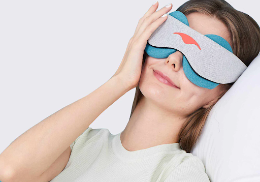 A girl wearing a light gray cooling eye mask touching the side of her face.