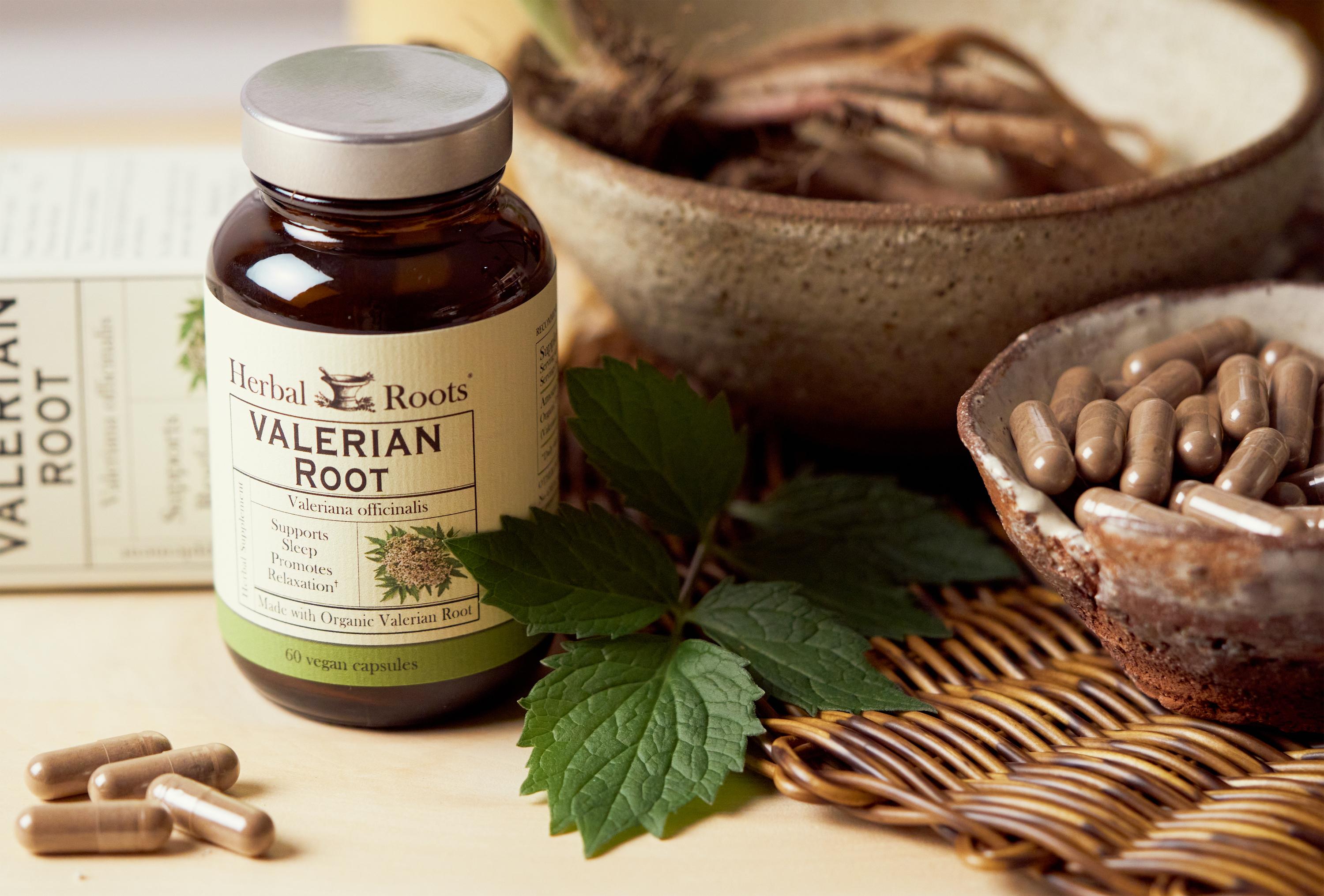Amber Bottle with silver lid, green and white label matching with box of Herbal Roots valerian root next to a flat wicker place mat with two bowls on it. The smaller bowl has valerian capsules in it and the larger bowl has valerian root in it. There are some capsules to the front left of the bottle.