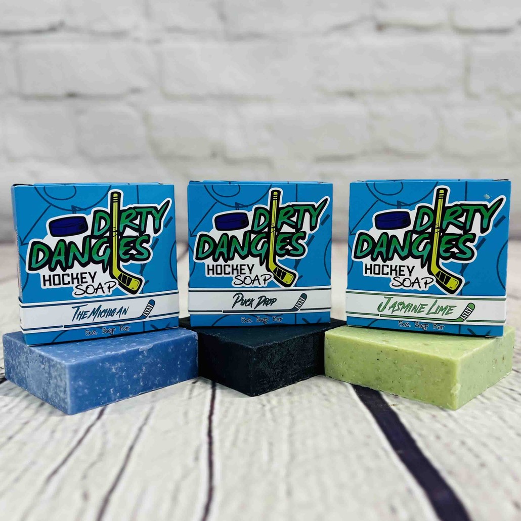 3 bars of dirty dangles hockey soap on a wood background. Blue, black and green. The michigan, puuck drop and jasmine lime.