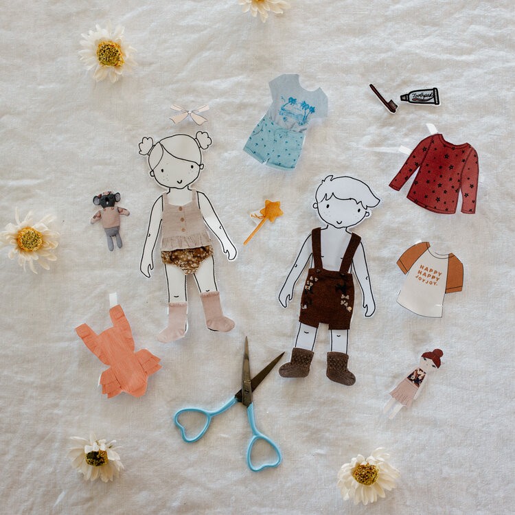 2 paper dolls with clothes lie on a bed with paper clothes surrounding them.
