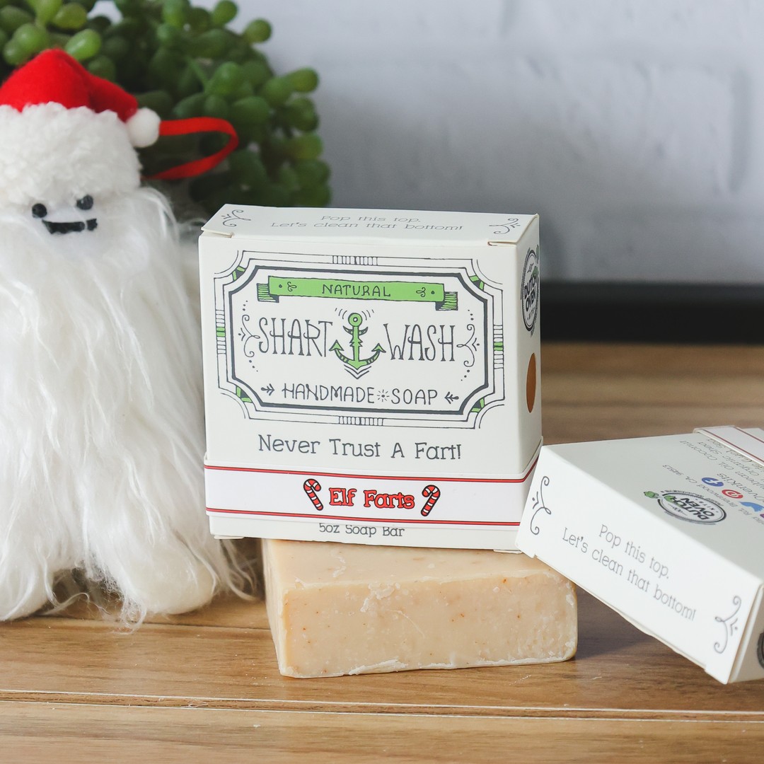 A tan bar of elf farts shart wash soap (Oatmeal spice scent) on a wood table with a christmas yeti