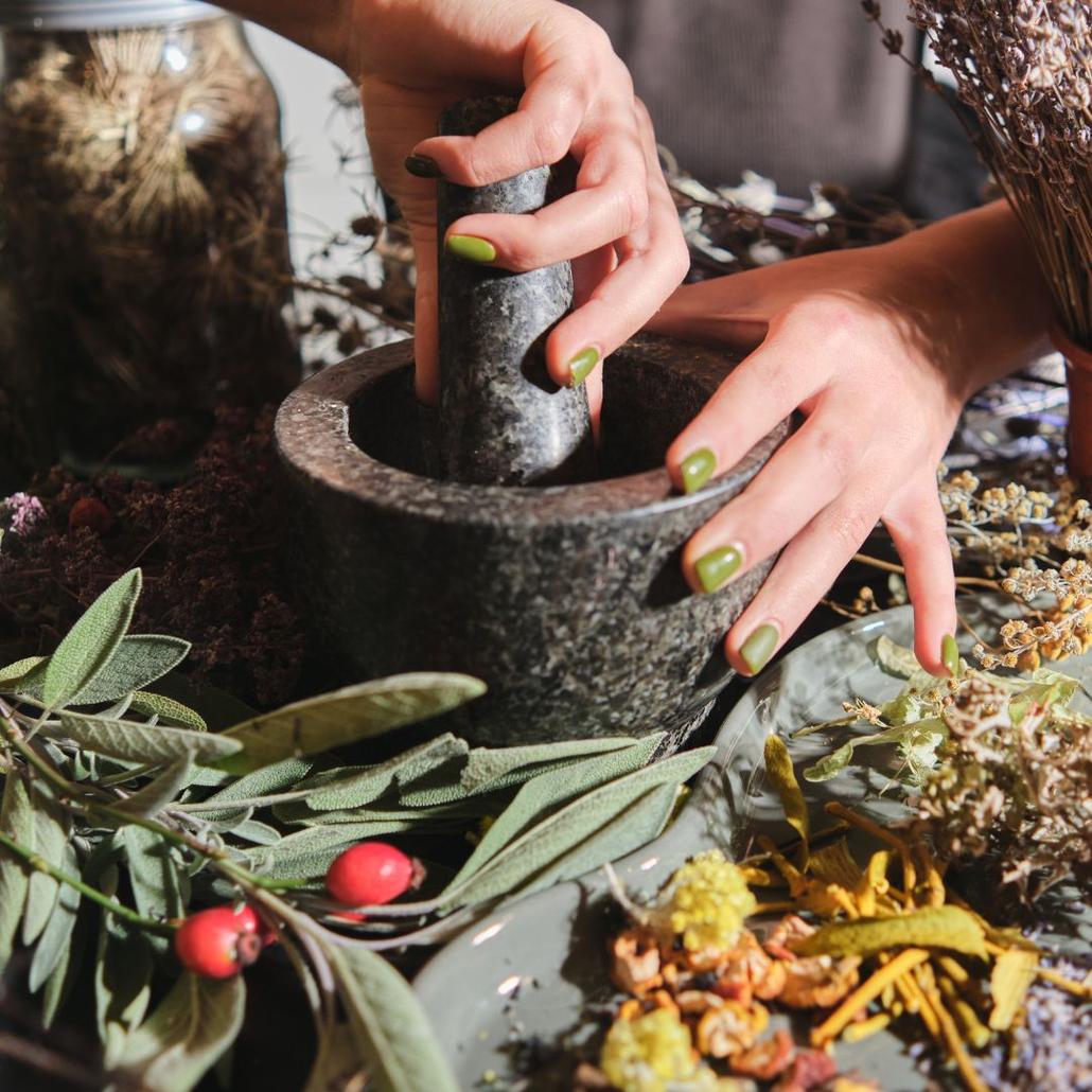 mortar and pestle being used by woman hands with various herbs surrounding it