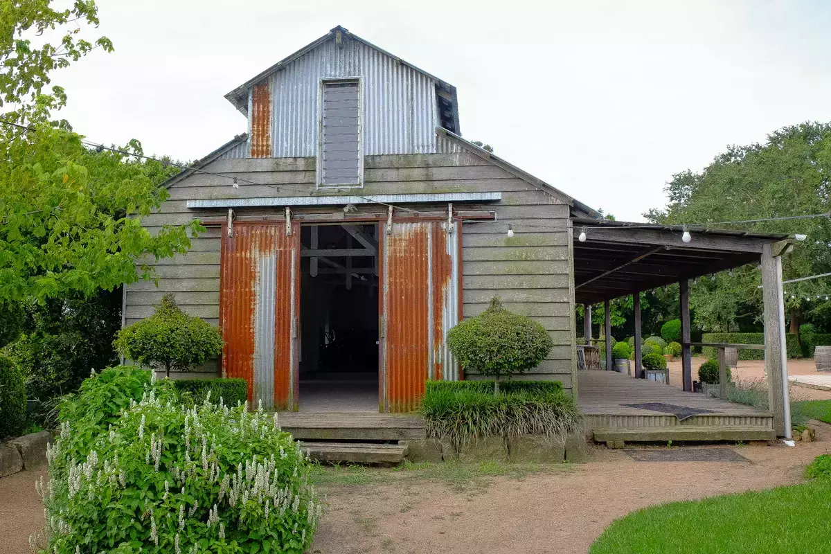 Rustic timber barn with weathered corrugated iron doors and siding, set within landscaped gardens, featuring a covered veranda and string lights for a countryside venue setting.