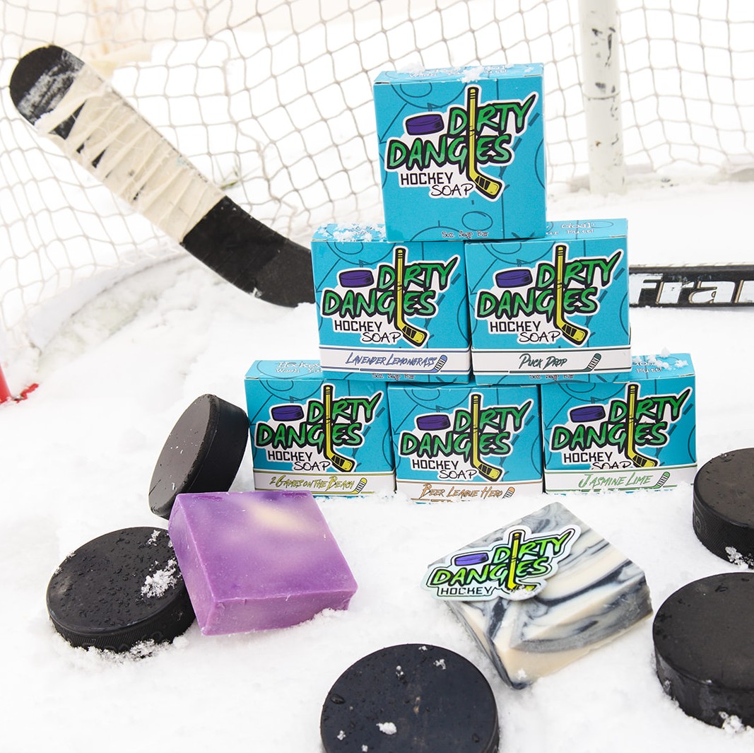 Bars of hockey soap sit in the snow with hockey pucks and a hockey stick. Dirty Dangles hockey soap.