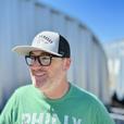 A man outside smiling wearing a light gray and black dirty dangles hockey co. mesh snapback trucker hat.