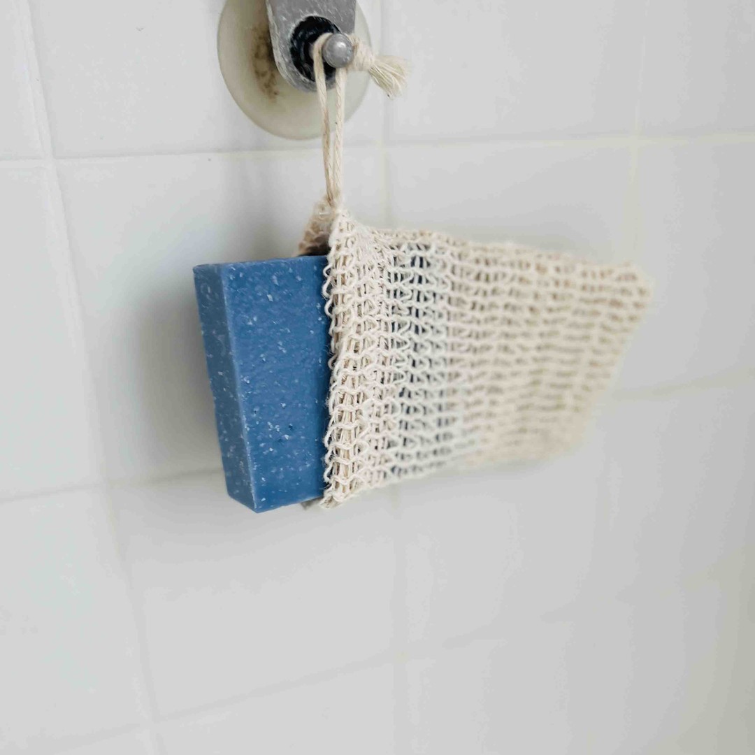 a tan sisal soap bag hangs in a shower with a blue soap bar inside