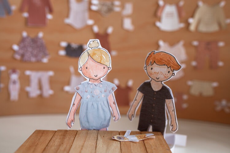 Two paper dolls stand wearing paper clothes with paper clothes hung up behind them.