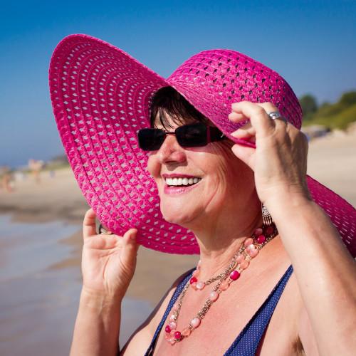 senior woman wearing a pink sun hat, while standing on the beach.