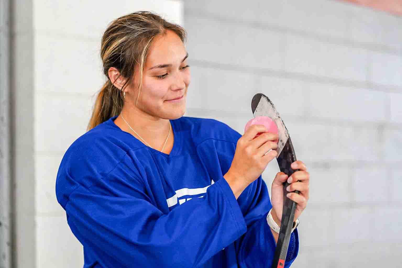 a female Hockey player applying pink stick wax to a hockey stick blade.