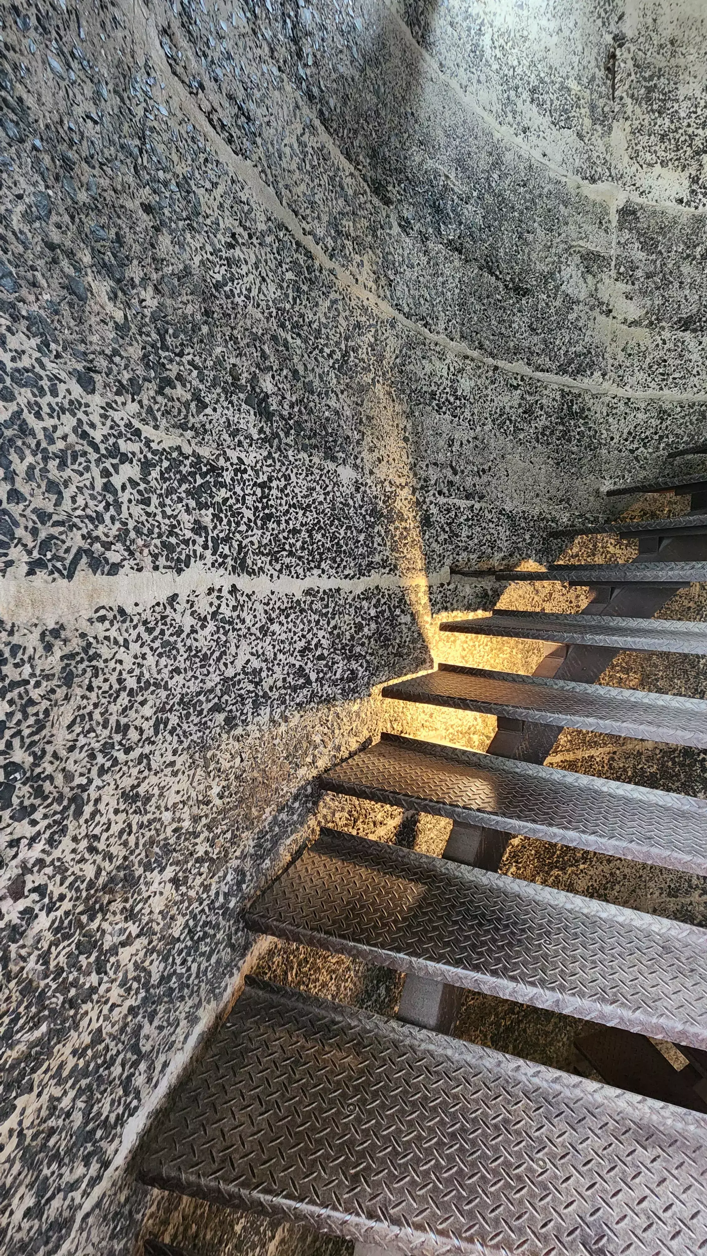Spiral metal staircase inside a concrete tower with textured aggregate walls and sunlight streaming through narrow openings.