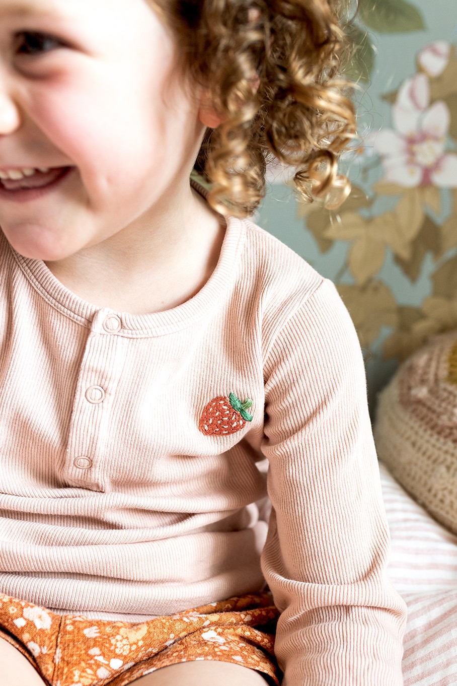 A little girl smiles, wearing a shirt with a strawberry embroidered on it.