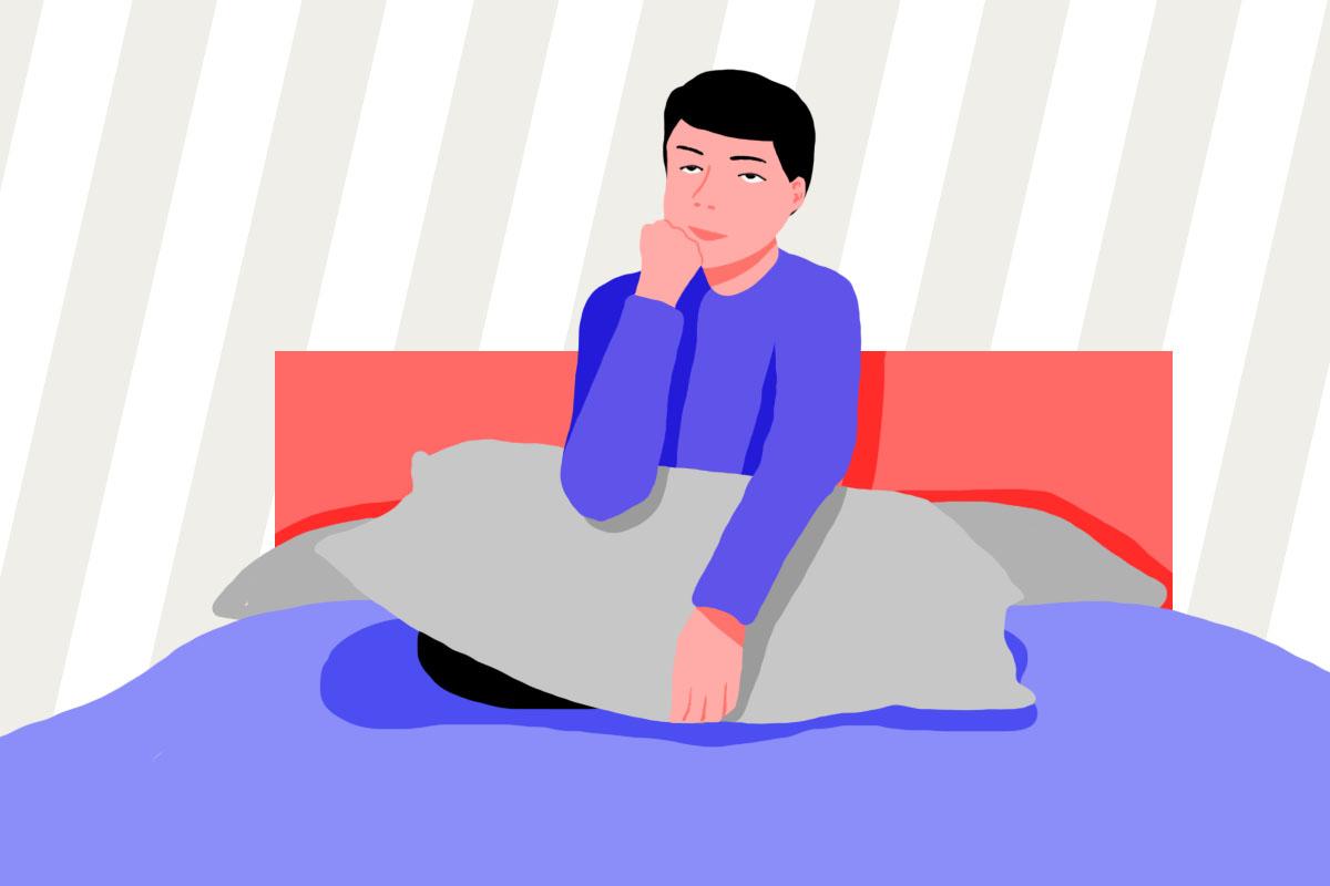 A man sitting cross-legged on a bed with a pillow resting on his knees, thinking about how many pillows you should sleep with.