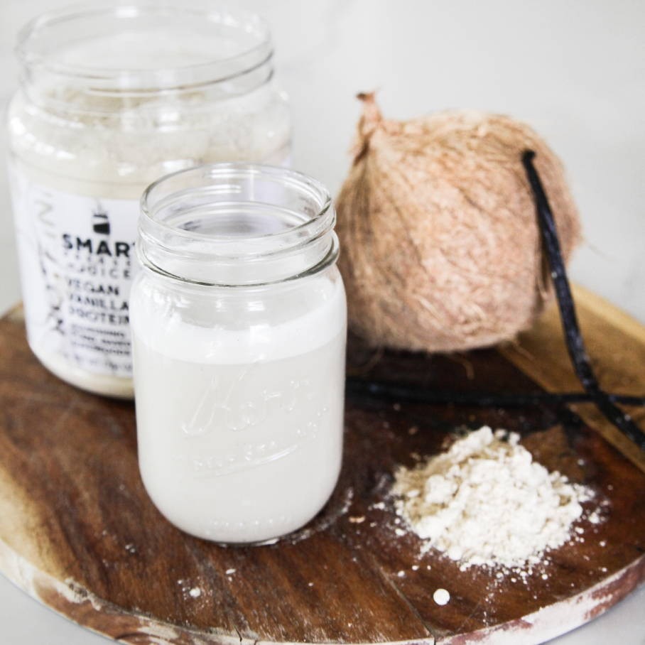 A mason jar with no lid filled with white smoothie set on a wooden circular board. Behind it is an open Vegan Vanilla Proteini jar on the left, and a whole coconut on the right. Next to it is is a pair of vanilla stems and a spoon of beige powder.