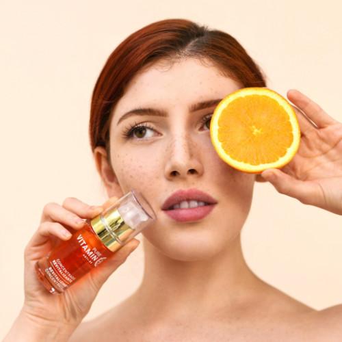 young model holding an orange slice in front of her right eye and holding up Noche's Vitamin C Serum in her other hand