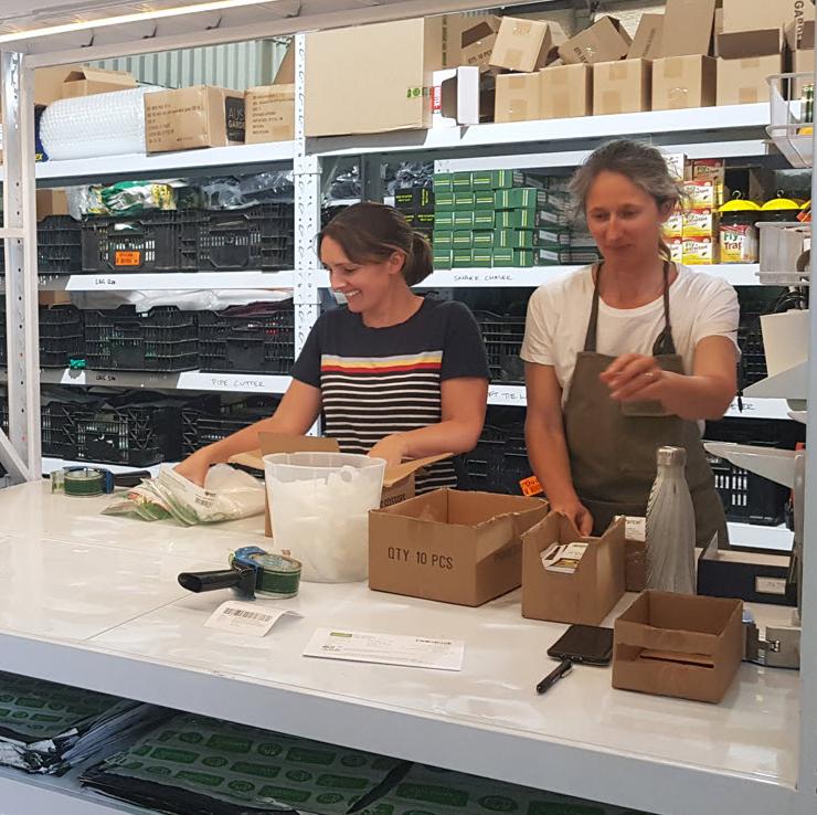 Two women packing gardening products at a workbench in a warehouse.