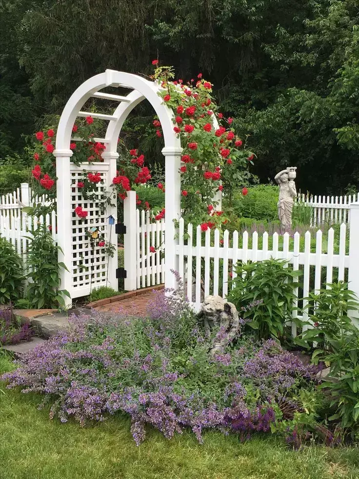 White garden arch and gate with red climbing roses and cottage garden plants.