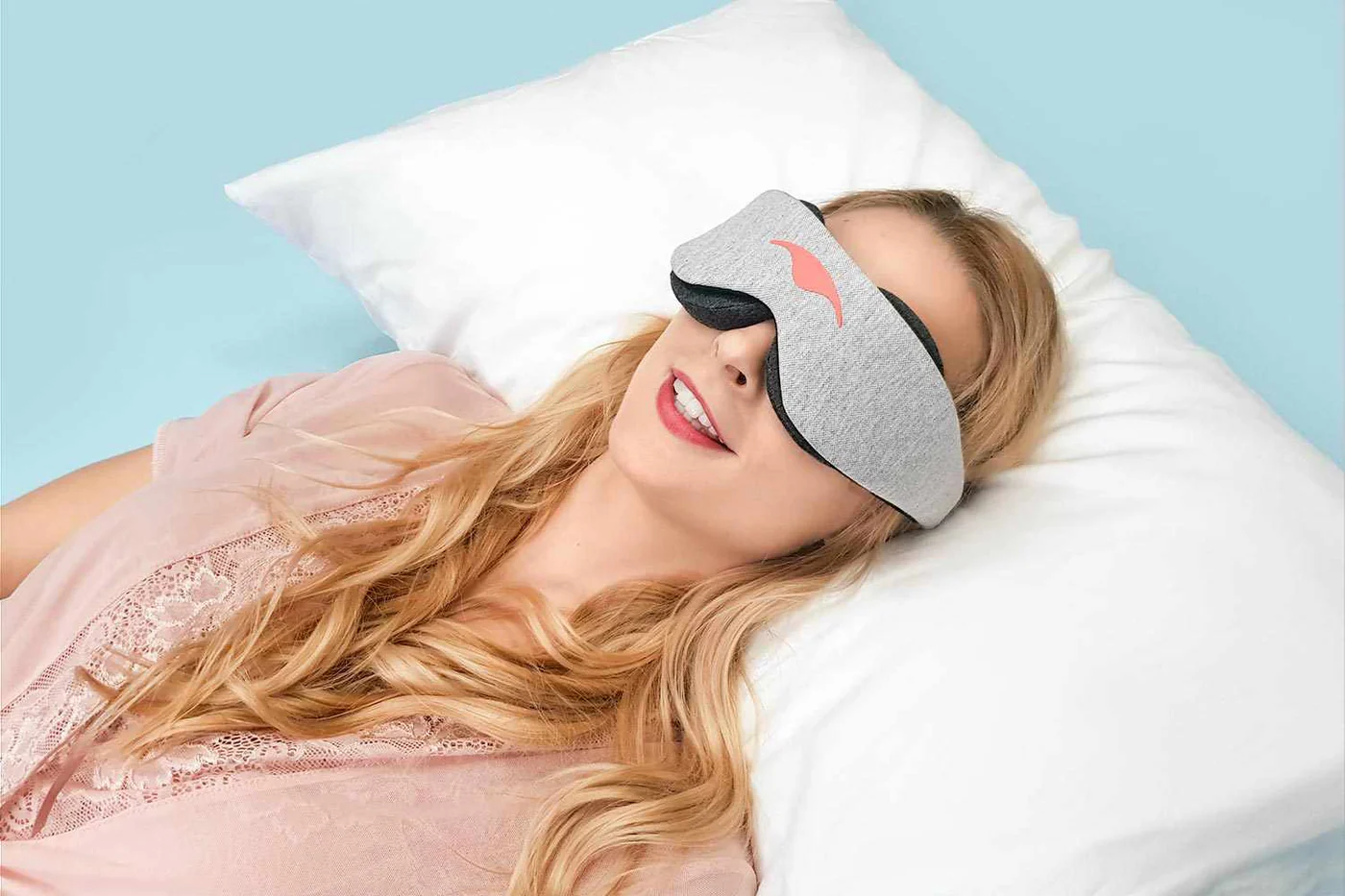A blonde girl lying down on a white pillow, wearing a gray sleep mask with eye cups.