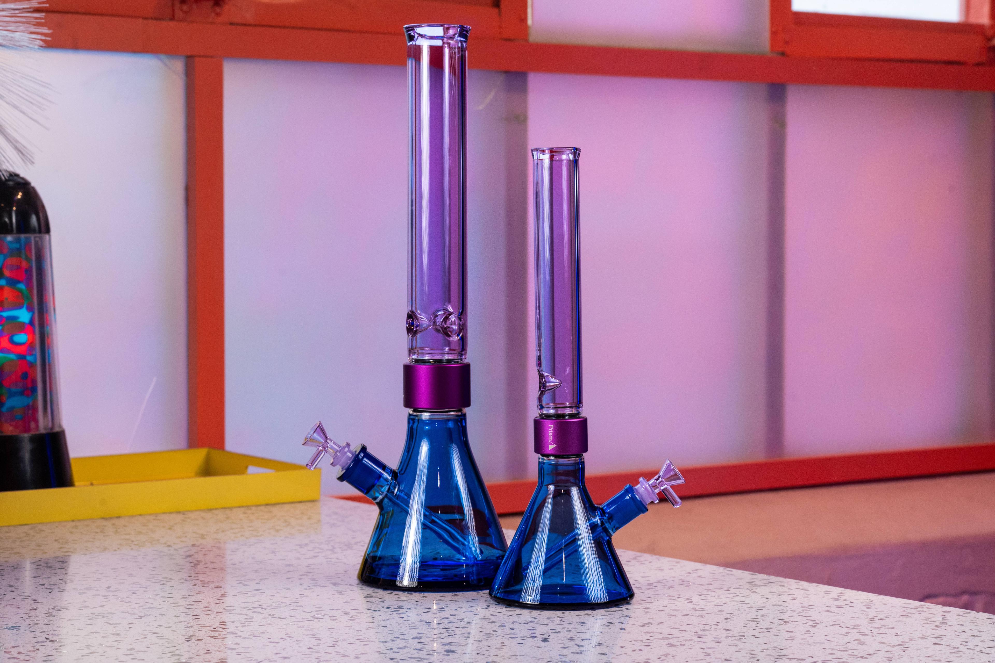 A side-by-side comparison of two blue and purple Prism modular glass bongs from the Duo Collection. On the left is the MINI size and on the right is the Classic size, both featuring tall Grape Jolly Rancher purple mouthpieces, purple Halo Connectors, and Sapphire Blue beaker bases.