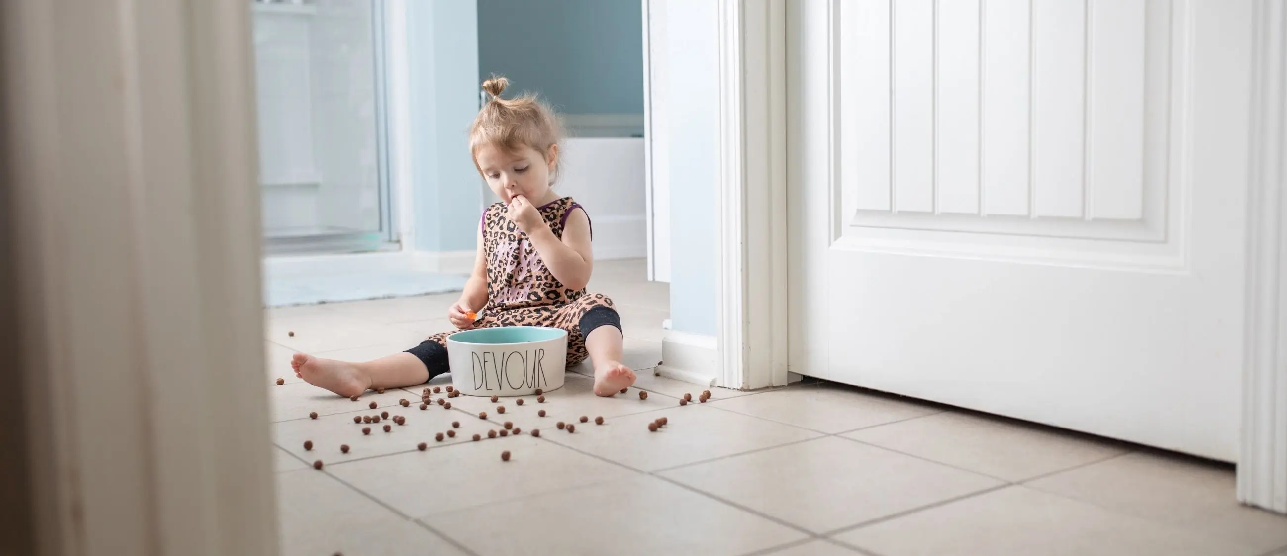 Toddler sitting on a tiled floor eating pieces of cat food from a bowl labeled "DEVOUR" with kibble scattered around, showing why it is important to keep a toddler out of the litter box and pet feeding areas.