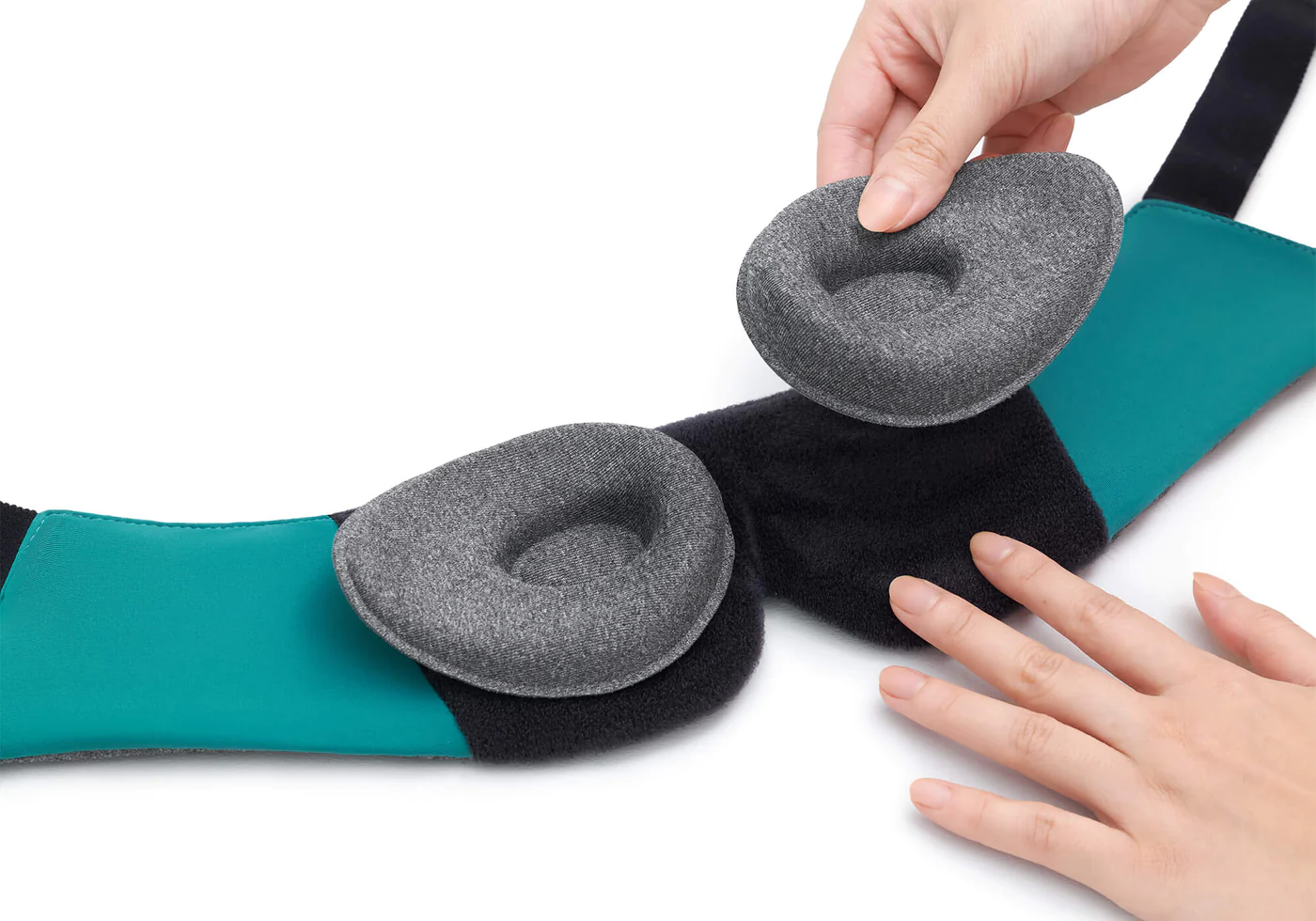 Hands attaching 1 of 2 gray tapered eye cups to the black and green interior of a sleep mask for headaches.