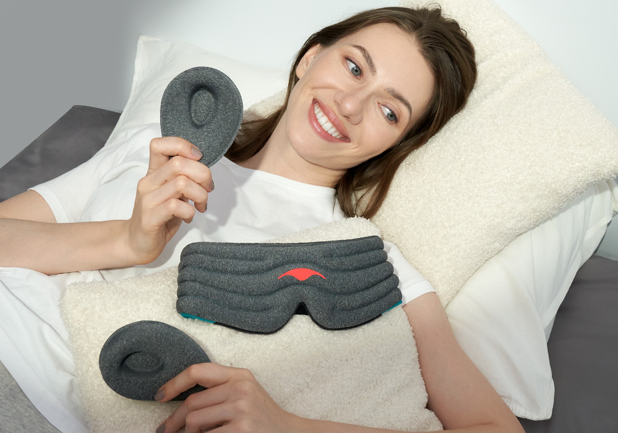 A smiling girl lying down on pillows with the gray head strap of the weighted eye mask lying on her front. She is holding a tapered eye cup in each hand.