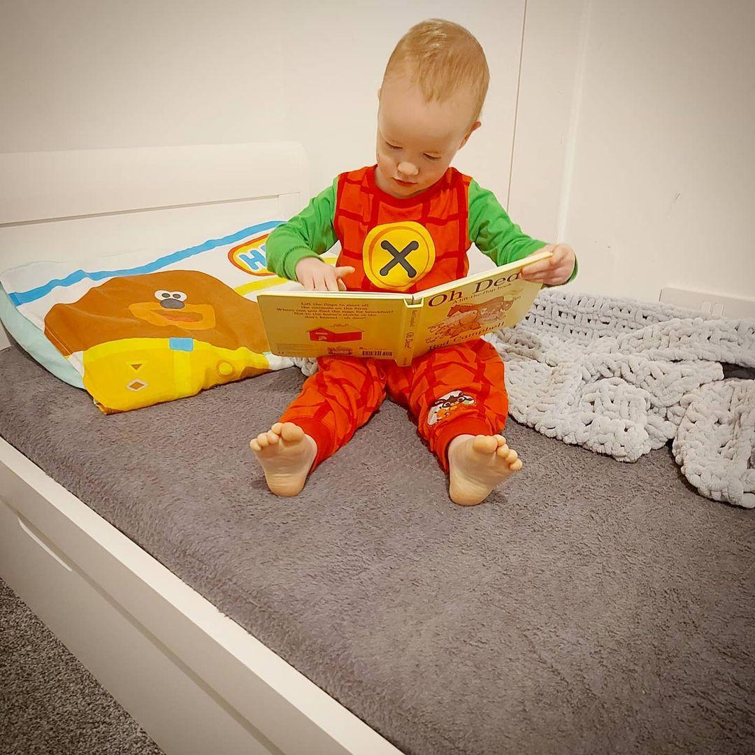Toddler boy reading a book in his bed. Sitting on large grey PeapodMat - 100% leakproof bed mat.