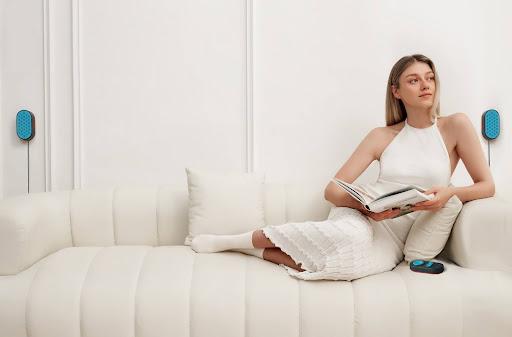 A woman reading on a couch while relaxing to sounds from the best white noise machine for sleeping. The device’s speakers are mounted on walls on either side of her. The control pad is beside her on the couch.